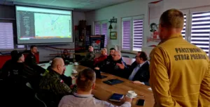 Strażak PSP odwrócony plecami, patrzy się w stronę sali konferencyjnej pełnej osób